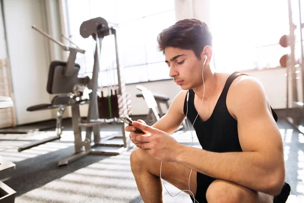 Hombre hispano en el gimnasio descansando, sosteniendo el teléfono inteligente, escuchando musi — Foto de Stock