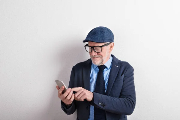 Smart senior man in suit, eyeglasses and cap, holding smartphone