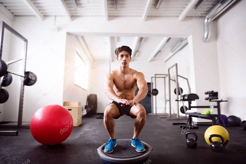 Young fit hispanic man in gym doing squats on fitness ball