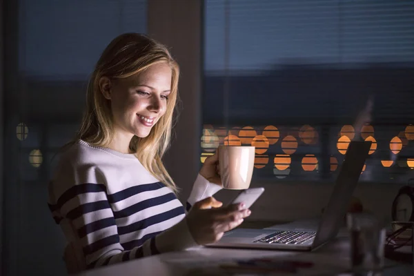 Женщина за столом, держит смартфон, работает на ноутбуке ночью . — стоковое фото