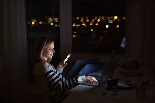 Mulher na mesa, segurando smartphone, trabalhando no laptop à noite . — Fotografia de Stock