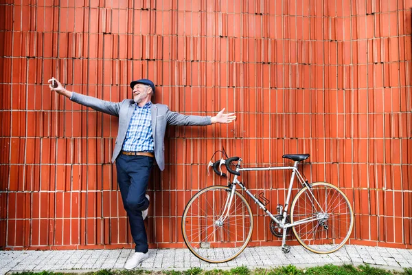 Hombre mayor con smartphone y bicicleta contra pared de ladrillo . — Foto de Stock