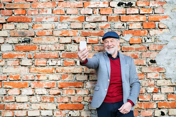 Hombre mayor con teléfono inteligente contra la pared de ladrillo tomando selfie . —  Fotos de Stock