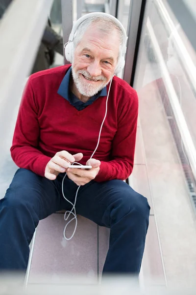 Uomo anziano con smartphone e cuffie seduto in passaggio . — Foto Stock