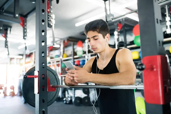 Homme hispanique dans la salle de gym au repos, tenant un téléphone intelligent, écoutant de la musique — Photo