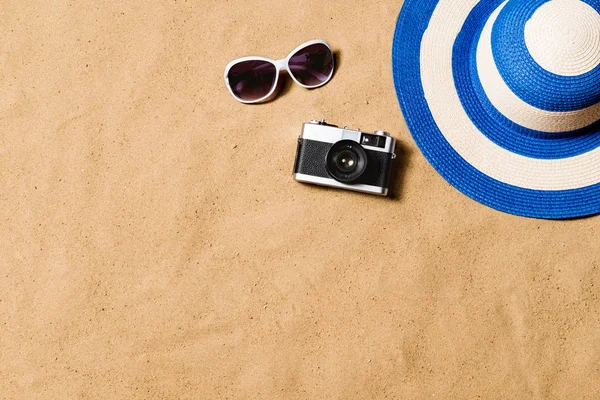 Sunglasses, wicker hat and camera. Sand background.