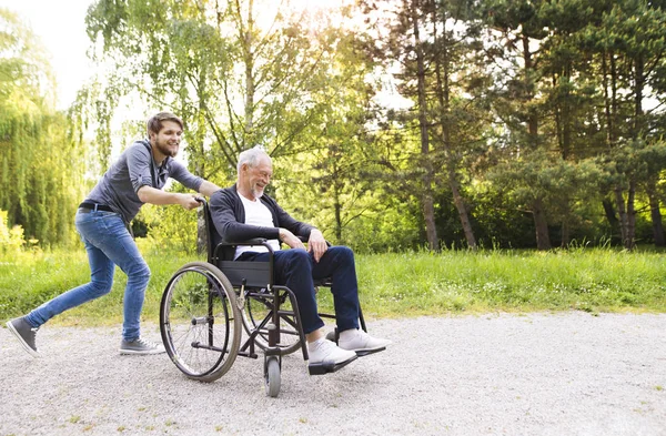 Hipster hijo corriendo con padre discapacitado en silla de ruedas en el parque . — Foto de Stock