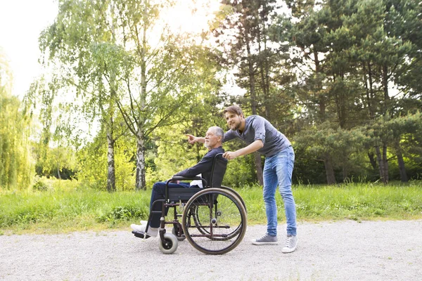 Hipster son walking with disabled father in wheelchair at park. — Stock Photo, Image