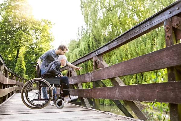 Hipster son walking with disabled father in wheelchair at park. — Stock Photo, Image