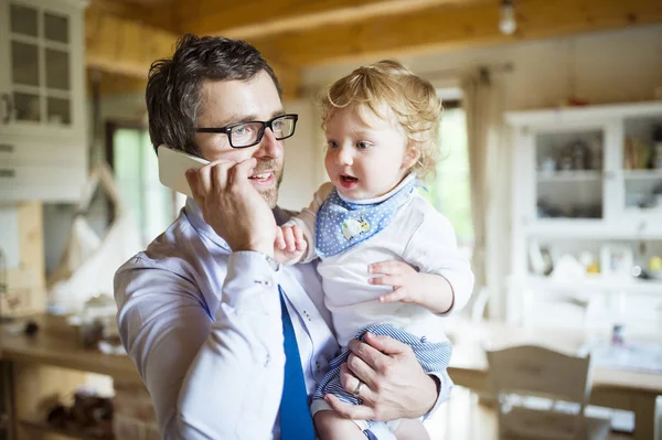 Επιχειρηματίας με έξυπνο κινητό τηλέφωνο στο σπίτι με το γιο στην αγκαλιά. — Φωτογραφία Αρχείου