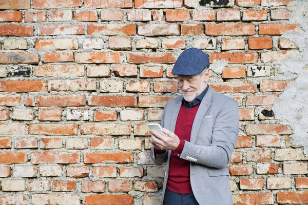 Uomo anziano con smartphone contro muro di mattoni, sms . — Foto Stock