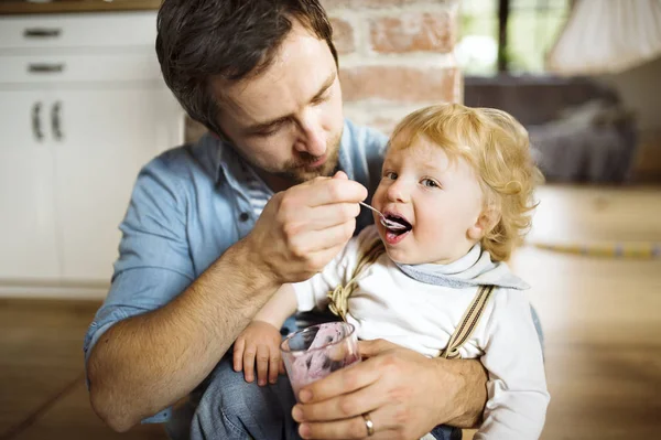 Padre joven en casa alimentando a su pequeño hijo con yogur . —  Fotos de Stock