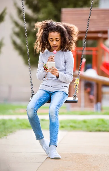 Africano menina americana no balanço com mensagens de texto telefone inteligente . — Fotografia de Stock