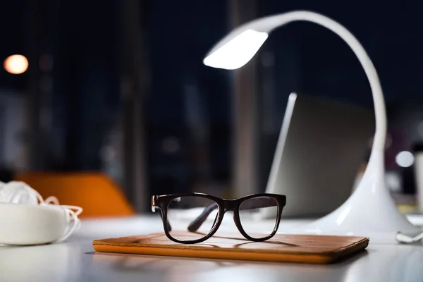 Office desk at night with eyeglasses, lamp and tablet.