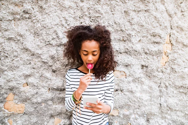 Bella ragazza afroamericana con smart phone, mangiare lecca-lecca — Foto Stock
