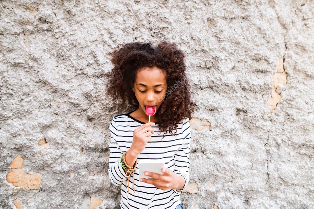 Beautiful african american girl with smart phone, eating lollipo