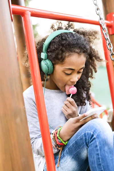 Africano menina americana no parque infantil com mensagens de texto telefone inteligente . — Fotografia de Stock