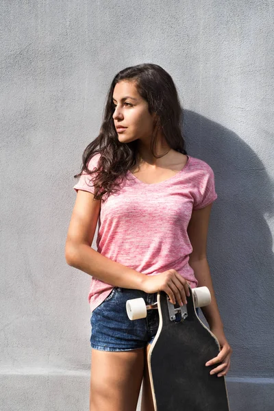 Krásná náctiletá studentka s skateboard proti betonové zdi. — Stock fotografie