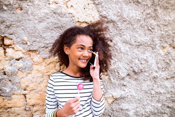 Menina americana africana bonita com telefone inteligente, comendo pirulito — Fotografia de Stock