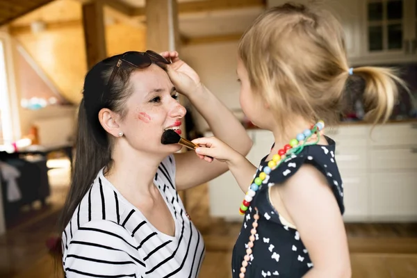 Menina bonito fazendo make-up para sua mãe atraente com escova . — Fotografia de Stock