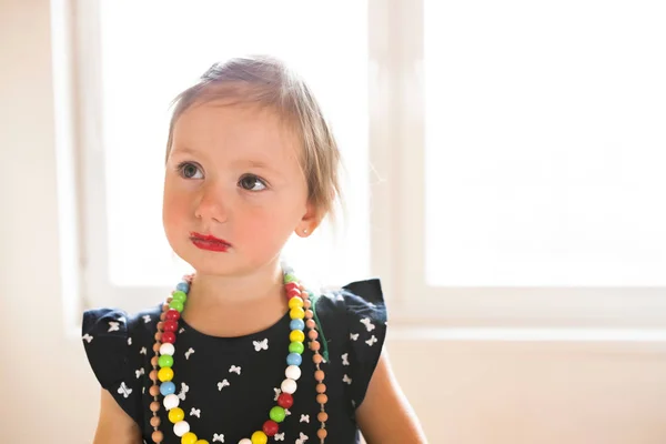 Carino bambina in abito a casa indossando rossetto rosso . — Foto Stock