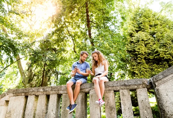 Young couple with smartphones in town sitting on concrete wall. — Stock Photo, Image