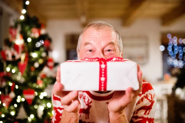 Senior man in front of Christmas tree holding a gift.