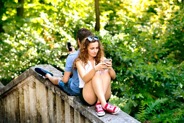 コンクリートの壁の上に座って町でスマート フォンを持つ若いカップルします。. — ストック写真