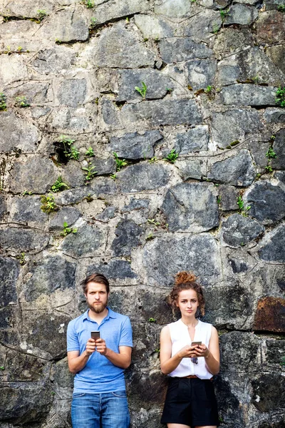 Young couple with smartphones against stone wall in town.