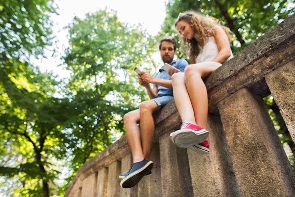 Young couple with smartphones in town sitting on concrete wall. — Stock Photo, Image