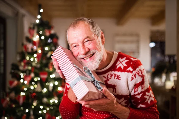 Senior man in front of Christmas tree holding a gift. — Stock Photo, Image