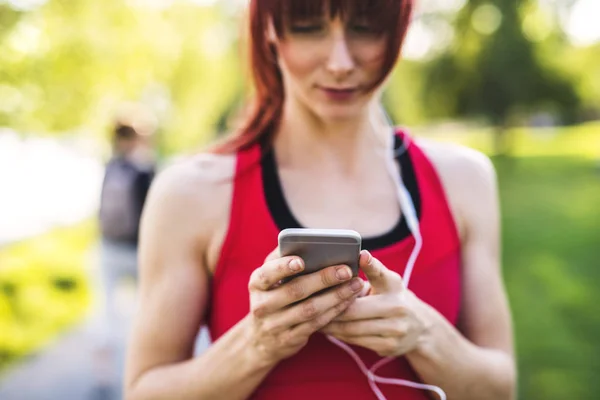 Junge Sportlerin im Park hält Smartphone in der Hand und hört Musik. — Stockfoto