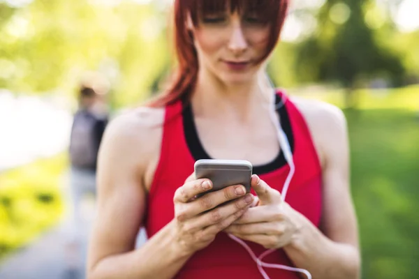 Jovem atleta no parque segurando telefone inteligente, ouvindo música . — Fotografia de Stock