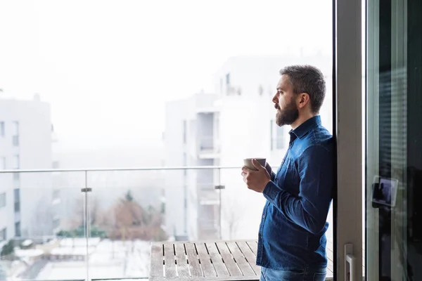Un uomo alla finestra che tiene una tazza di caffè a casa . — Foto Stock