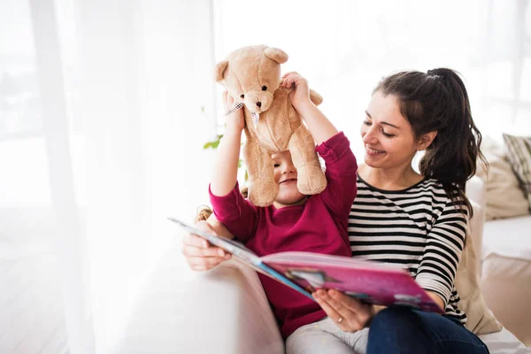 Madre joven y una niña pequeña con oso de peluche en casa, leyendo un libro . —  Fotos de Stock