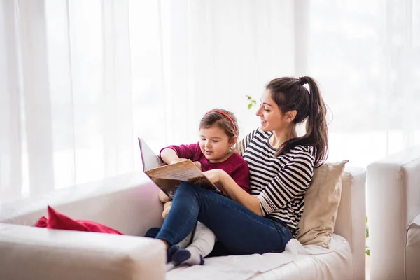 Jonge moeder met een klein meisje thuis, het lezen van een boek. — Stockfoto