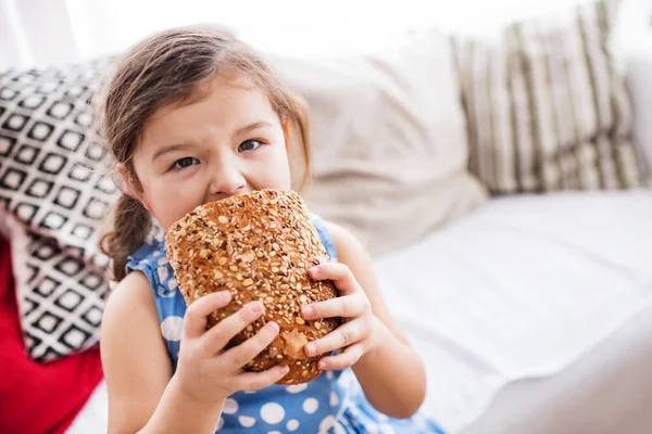 Uma menina pequena em casa comendo um pão . — Fotografia de Stock