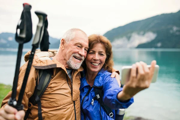 Un couple de retraités debout au bord du lac dans la nature, prenant selfie . — Photo