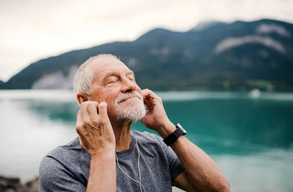 Starší muž se sluchátky stojící u jezera v přírodě, poslouchající hudbu. — Stock fotografie