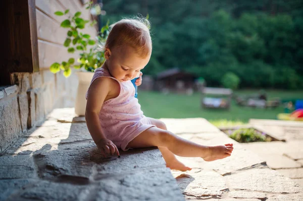 Een schattig peuter meisje zitten buiten in de voorkant van huis in de zomer. — Stockfoto