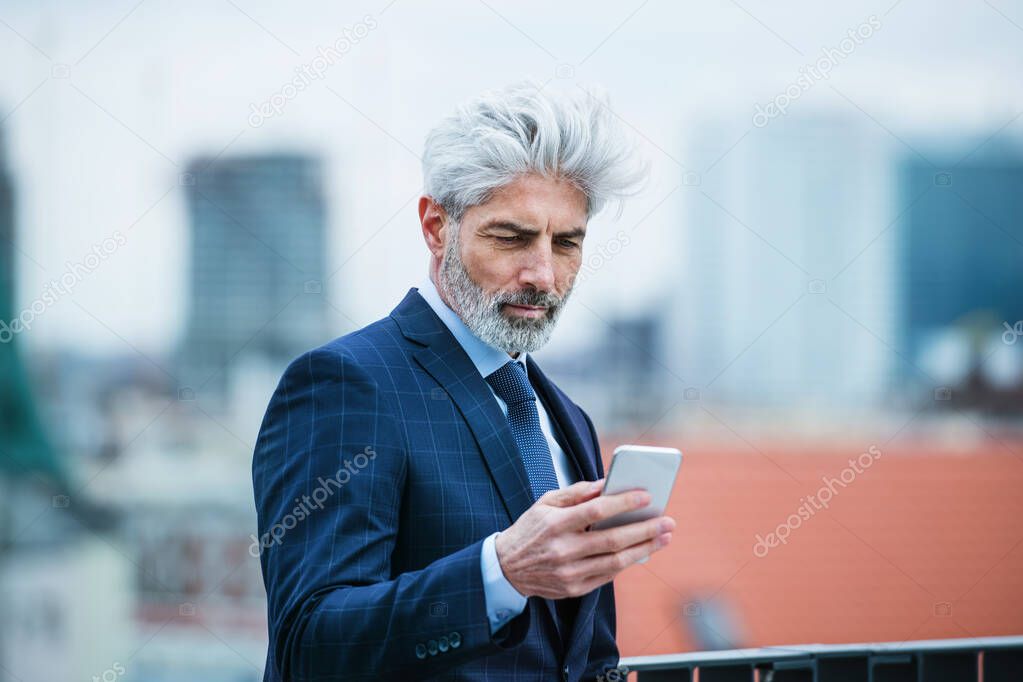 A mature businessman standing on a terrace, using smartphone.