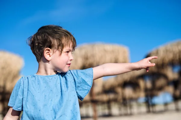 Una niña pequeña parada entre sombrillas de paja al aire libre en la playa . — Foto de Stock