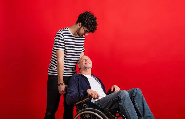 Young man and senior father in wheelchair in a studio on red background. — Stock Photo, Image