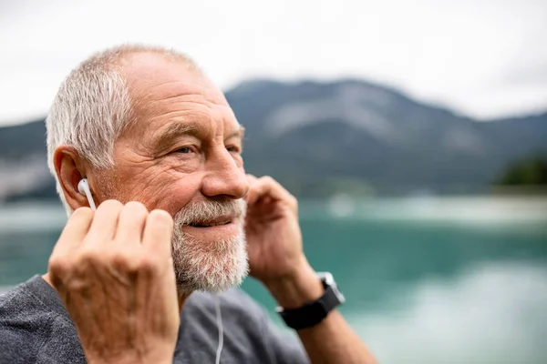 Hombre mayor con auriculares de pie junto al lago en la naturaleza, escuchando música . — Foto de Stock