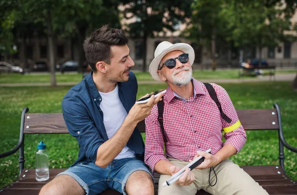 Young man and blind senior sitting on bench in park in city, using smartphone.