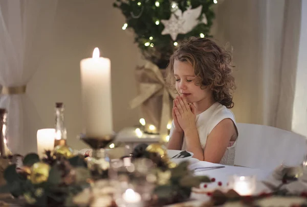Liten flicka sitter vid bordet inomhus vid jul, be. — Stockfoto