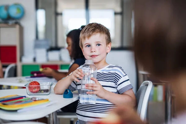 Small school boy sitting at the desk in classroom, drinking water. — Stock Photo, Image