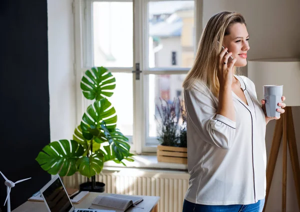 Jeune femme avec smartphone debout à l'intérieur dans le bureau à la maison, travaillant . — Photo