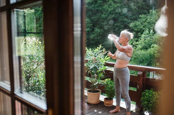 Una mujer mayor con sujetador deportivo al aire libre en una terraza en verano, agua potable . — Foto de Stock