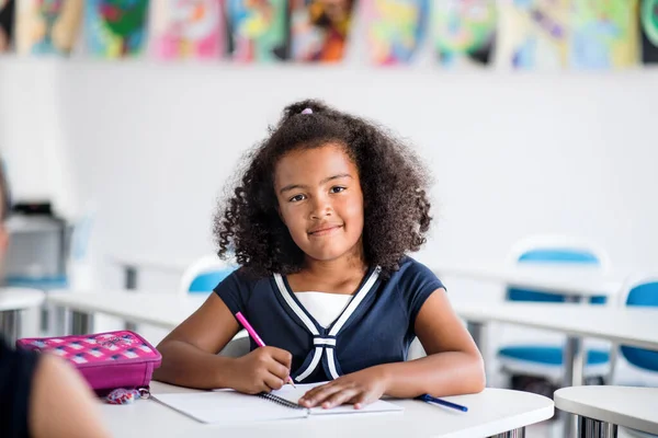 Маленькая школьница, сидящая за столом в классе, пишет: . — стоковое фото